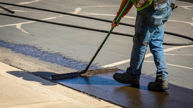 Worker brushing black sealcoat across a parking lot